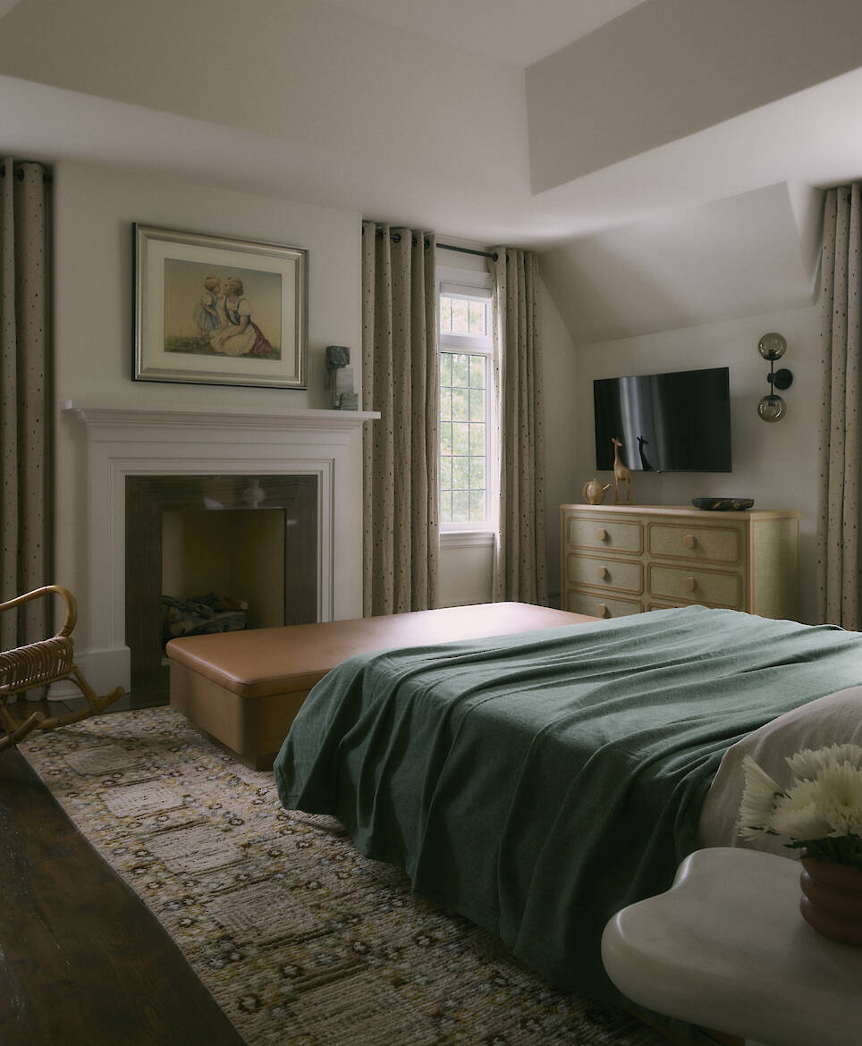 This inviting bedroom, part of a recent Toronto home renovation, features a green bedspread, fireplace, ottoman, dresser with a TV, armchair, and window with drapes. A stylish r