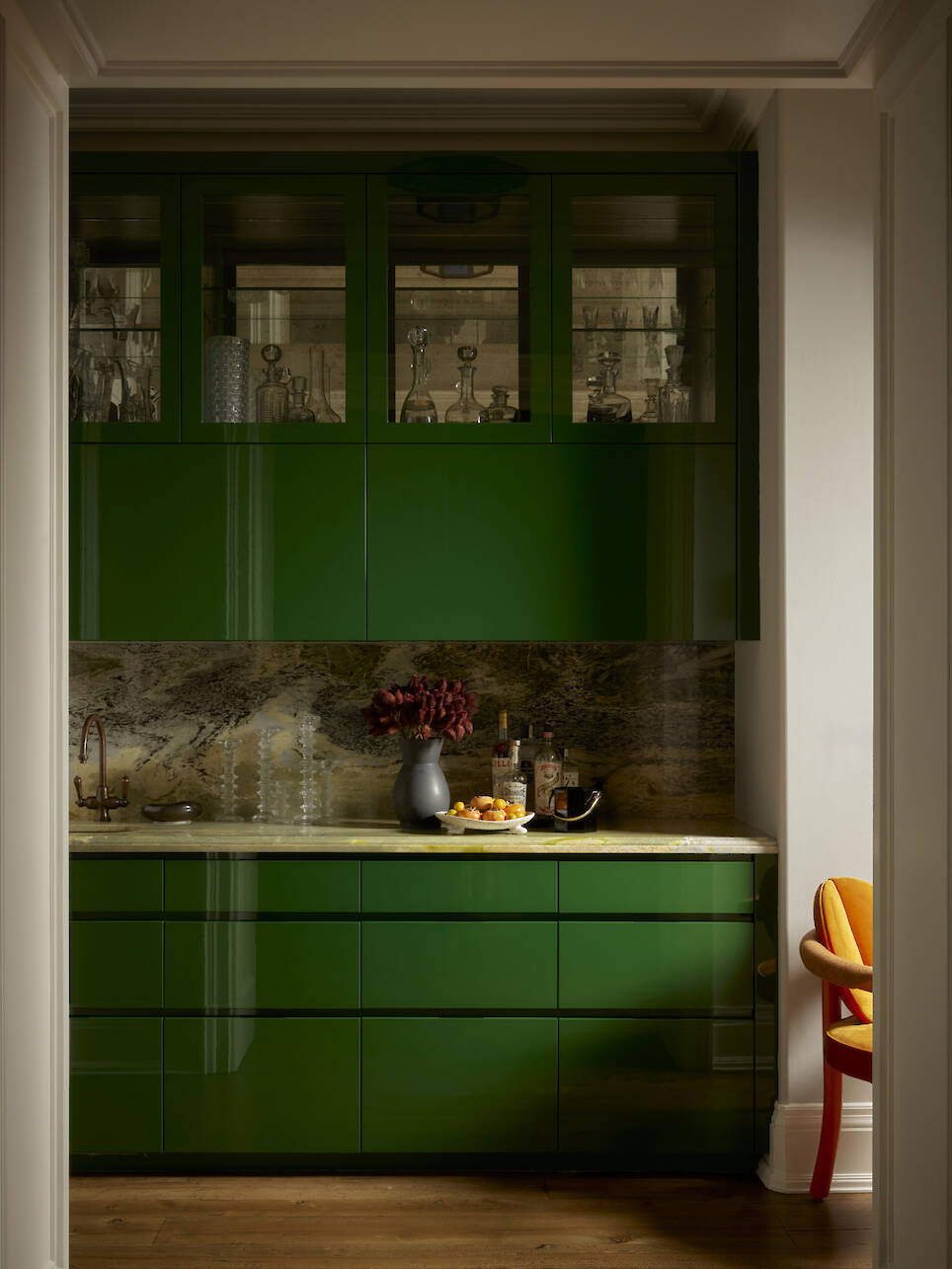 Small kitchen with glossy green cabinets, marble countertop and backsplash, glassware in upper shelves, and a vase with red flowers next to bottles and a bowl of lemons on the c