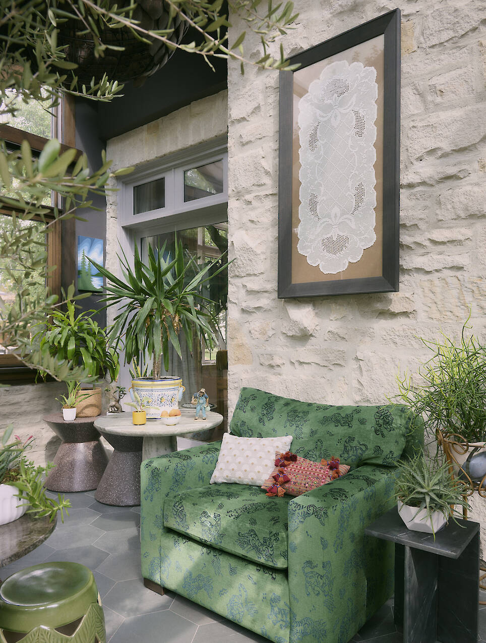 Green armchair with patterned pillow sits near potted plants, small tables, and a stone wall with a framed lace artwork in a sunlit indoor space—a cozy corner that beautifully