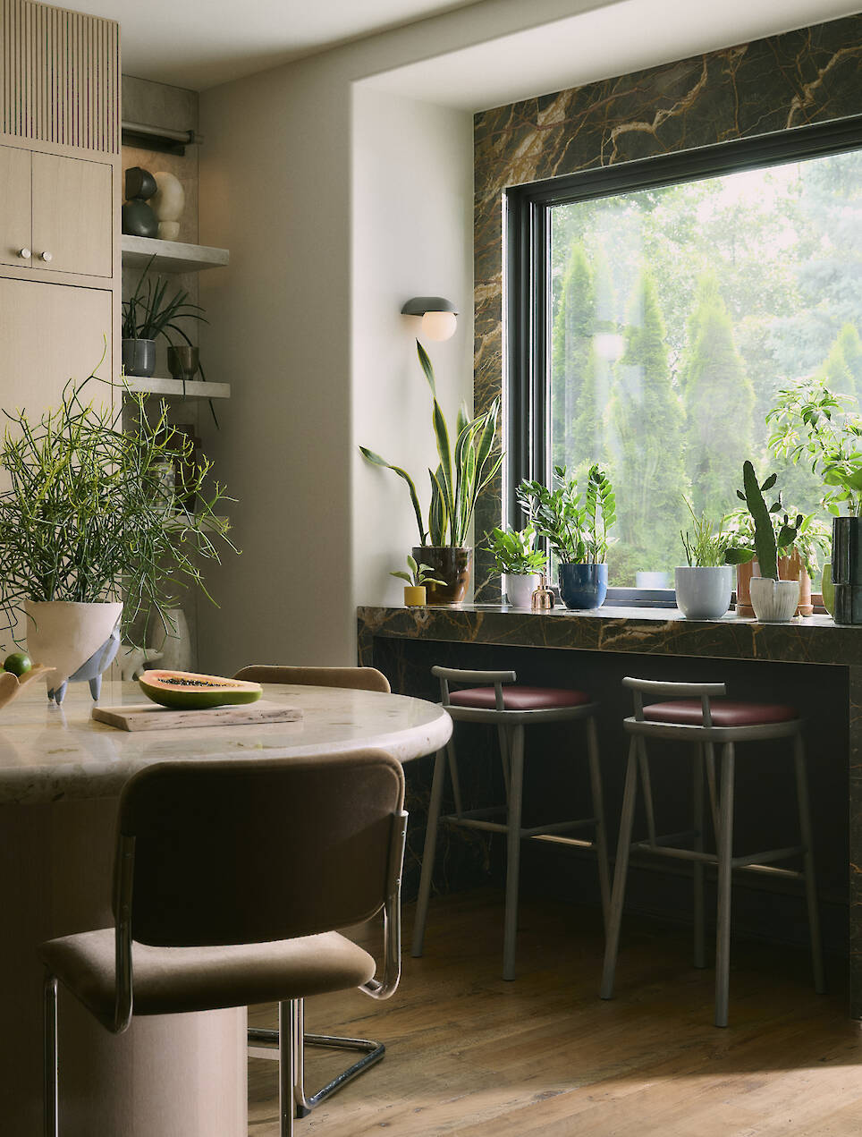 A modern kitchen, recently transformed as part of a Toronto home renovation, features a marble table, two chairs by a large window, and various potted plants on the windowsill a