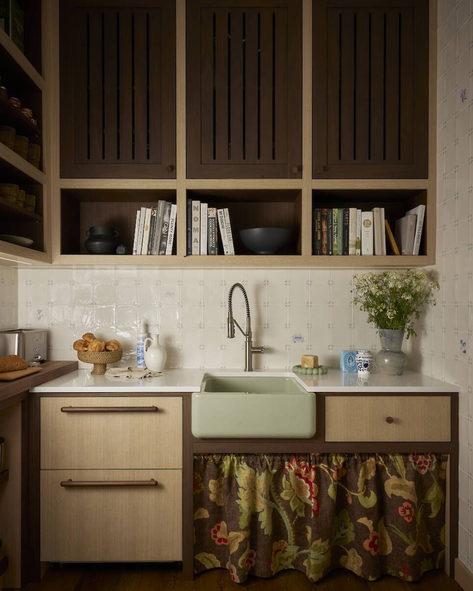 Small kitchen with open shelves, a green farmhouse sink, floral curtain under the sink, upper wooden cabinets, and books and dishes on the counter and shelves—charming touches