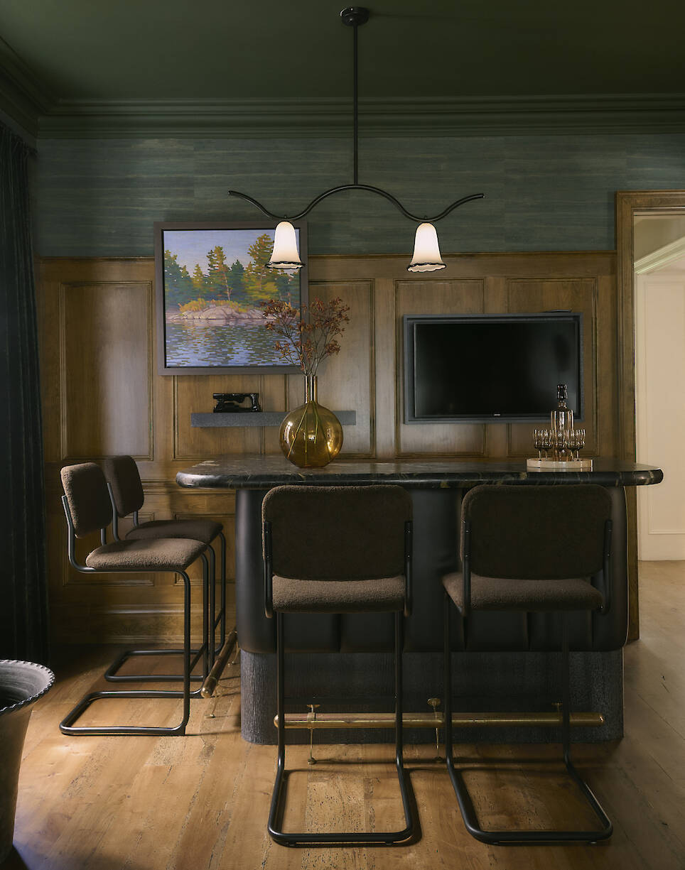 Home bar area with three barstools, a dark counter, wall-mounted TV, framed lake painting, and a glass vase with branches on the counter in a wood-paneled room—an inviting spa