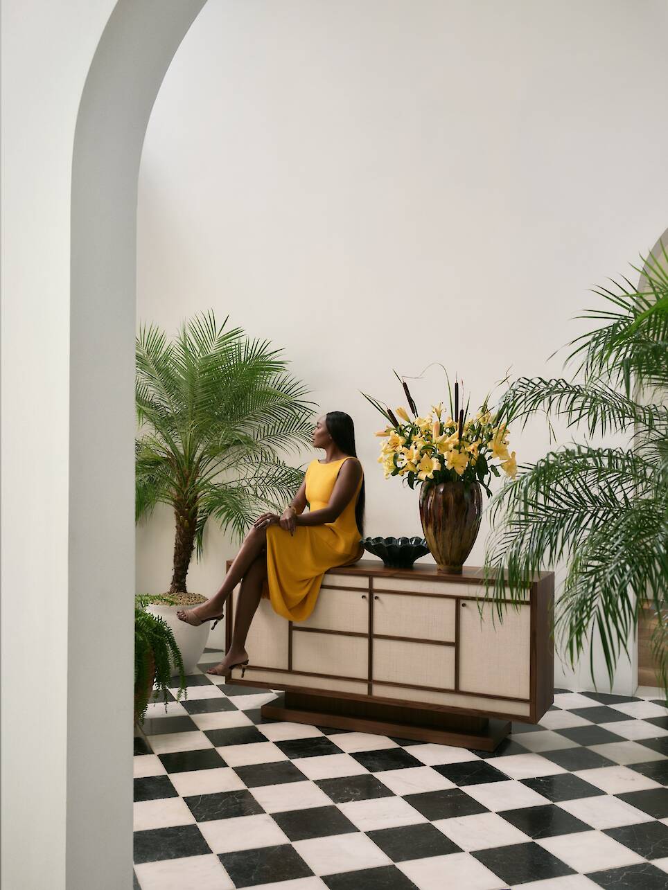 A woman in a yellow dress sits on a Crate & Barrel sideboard in a room with black and white checkered floor, potted plants, and a tall flower arrangement—a setting reminiscent