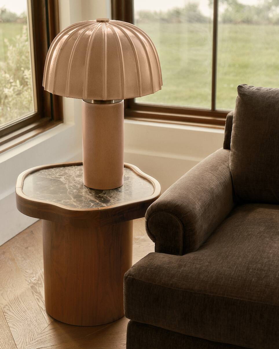A bronze-colored lamp with a dome-shaped shade, reminiscent of the elegant style found in the Venus Williams Crate & Barrel collection, sits on a marble-top wooden side table ne