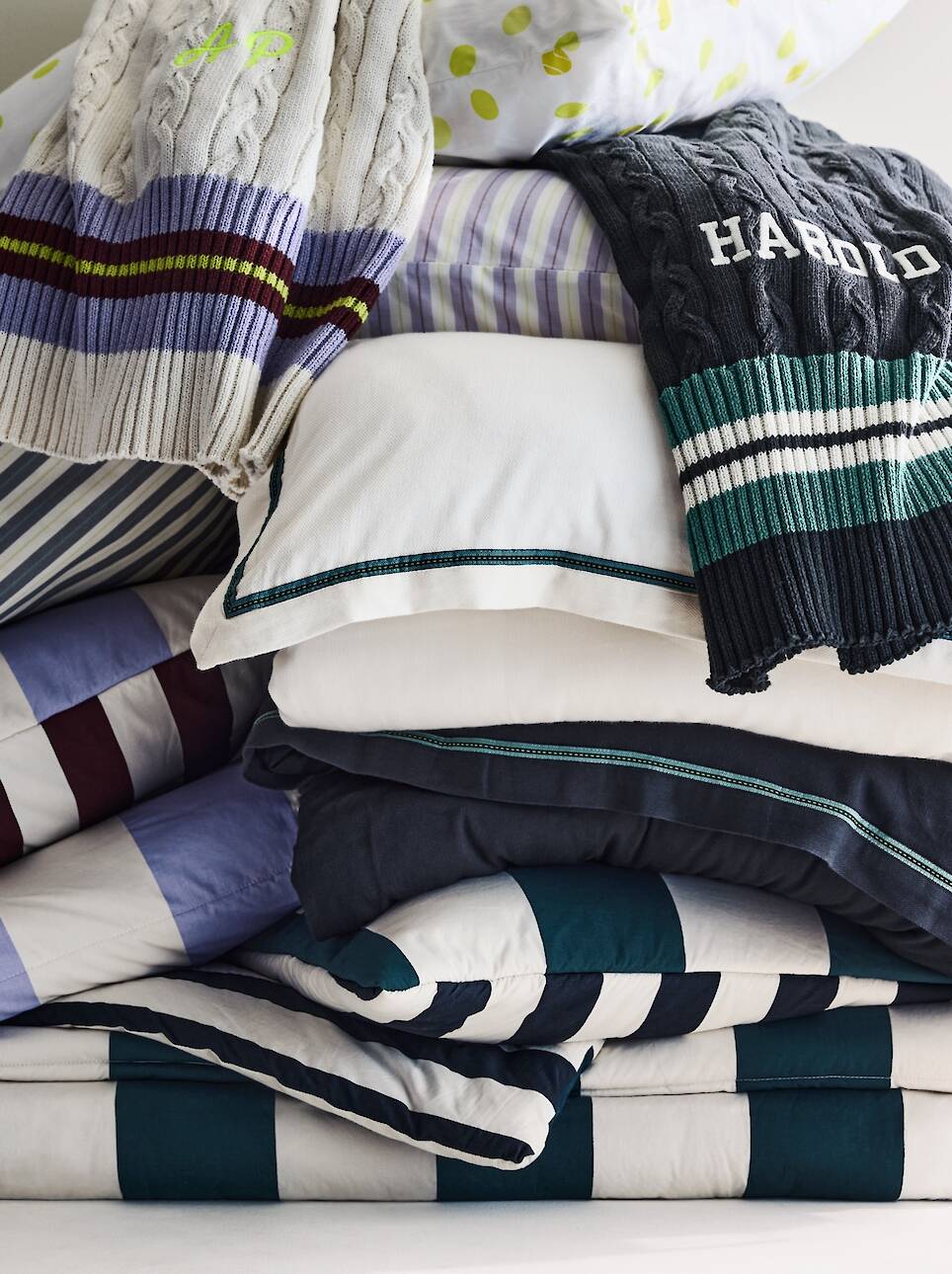 Stack of neatly folded bedding with striped patterns and pillowcases, topped with two sweaters draped over the pile, brings to mind the sophisticated comfort found in the Venus
