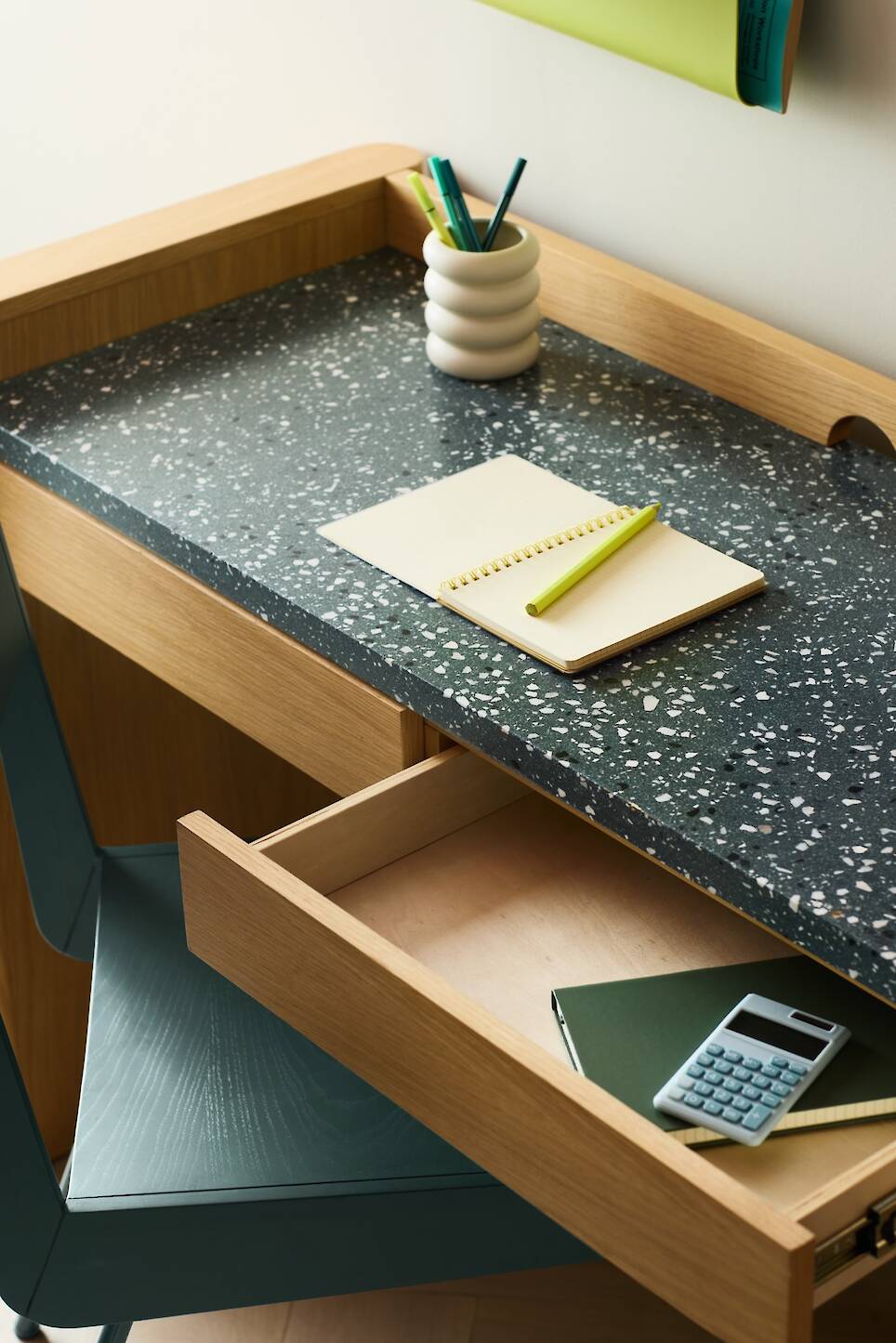 A terrazzo desk with an open drawer, a notebook and pen on top, a vase with pens, and a calculator and notebook inside the drawer—an elegant workspace inspired by Venus Willia