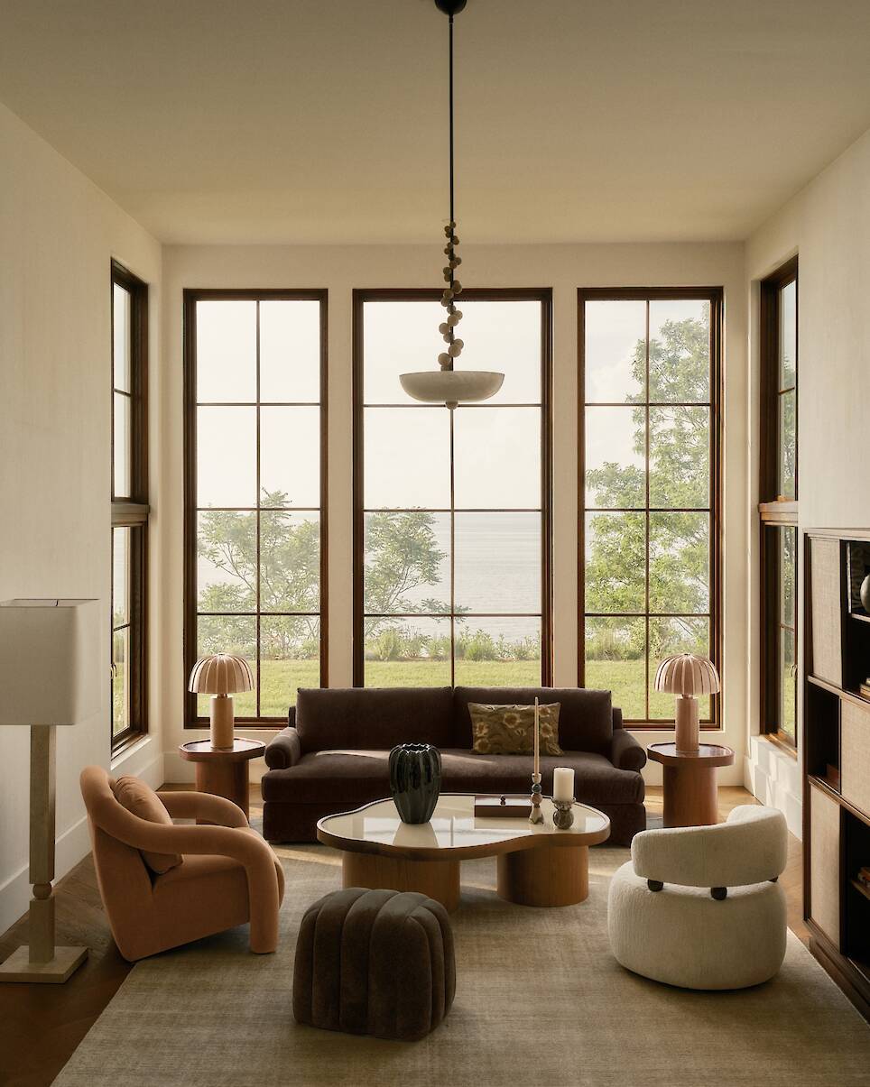 Modern living room with large windows, neutral furniture, three chairs, a sofa, coffee table, table lamps, and a serene view of trees and water outside. The space is elegantly s