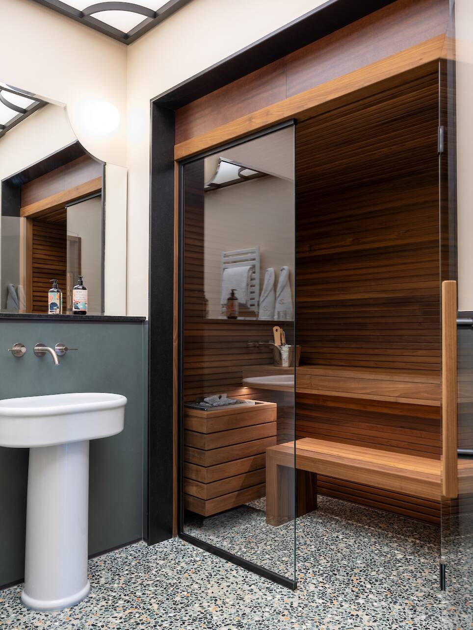 Modern bathroom with a pedestal sink, wall mirror, terrazzo floor, and a wooden sauna with a glass door and bench—echoing the refined aesthetic of Elizabeth Unique Venice. Tow