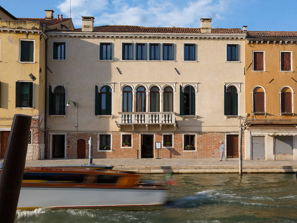 A boat moves along a canal in front of a beige historic building with arched windows and a balcony, capturing the timeless charm of Venice, Italy—an atmosphere reminiscent of