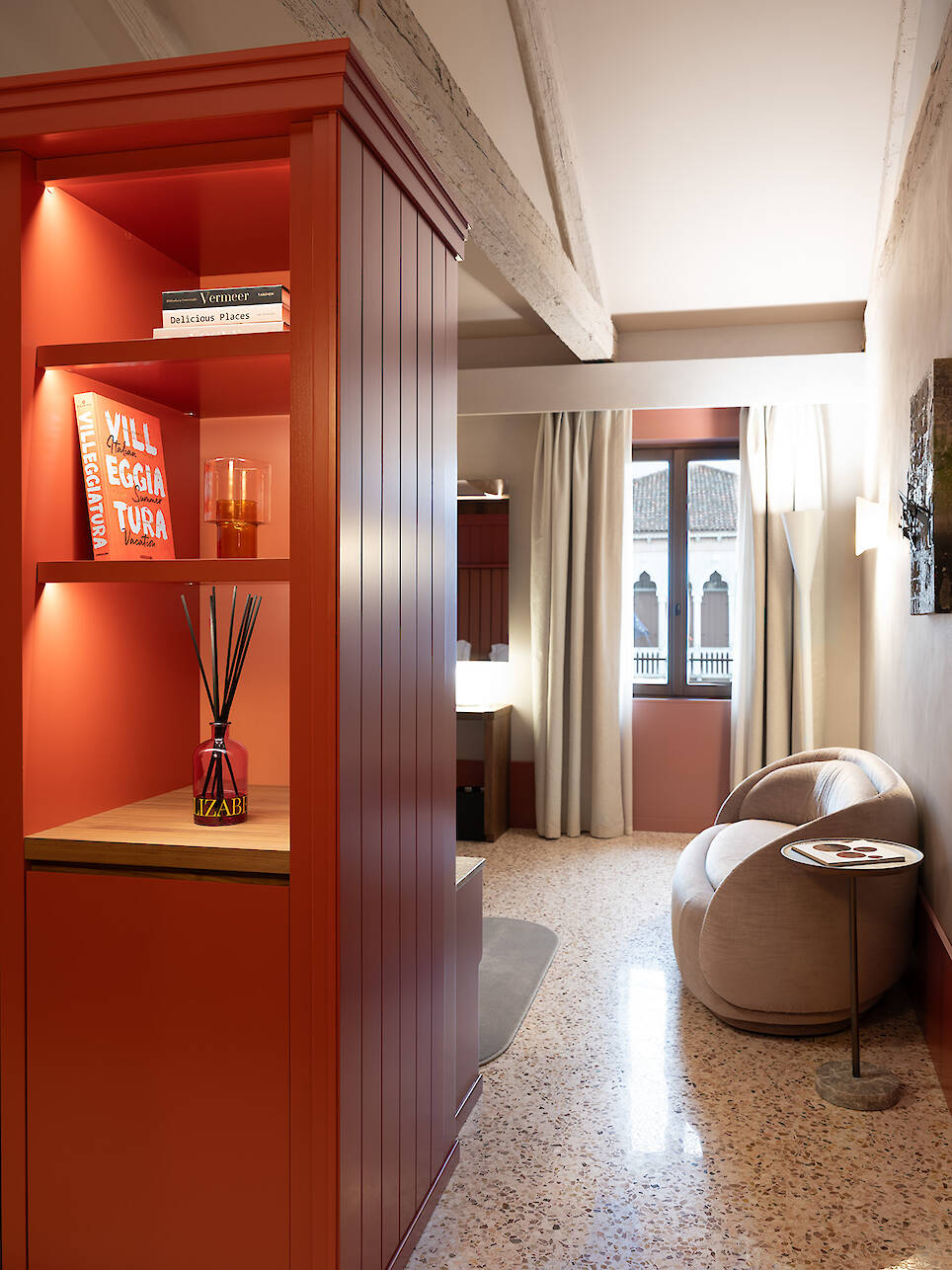A modern room inspired by the elegance of Elizabeth Unique Venice features a red shelving unit with books and decor, a round beige chair, a small side table, and a window with w