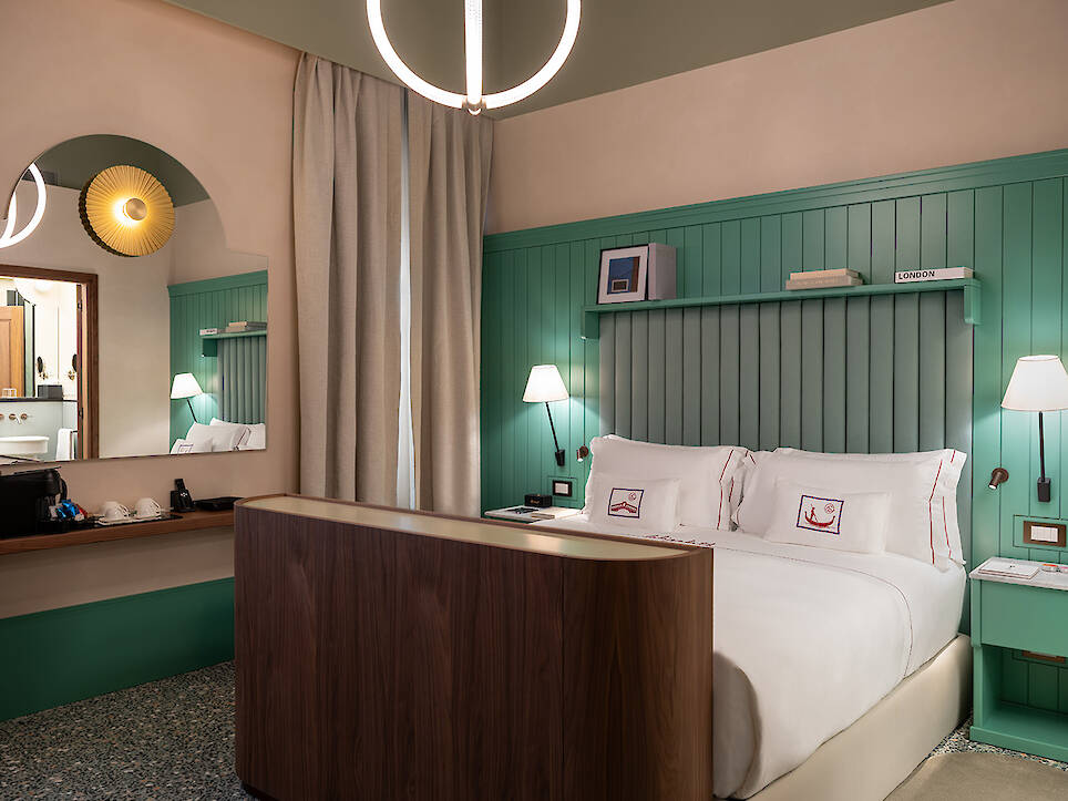 Hotel room with green paneled walls, double bed with white linens, a wooden console at the foot of the bed, wall lamps, a mirror, a curtain, and a round ceiling light—an invit