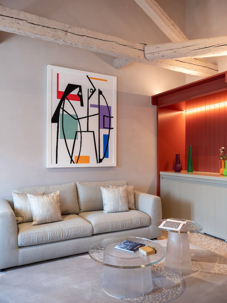 A modern living room with a beige sofa, abstract art on the wall, a glass coffee table, and a red alcove with vases evokes the sophisticated atmosphere of the Elizabeth Unique V