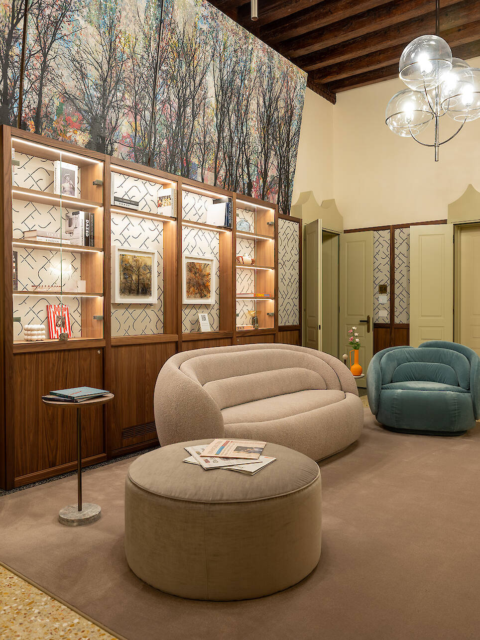 Modern lounge inspired by the sophisticated charm of Elizabeth Unique Venice, featuring a beige sofa, blue armchair, round ottoman, wall shelving, and wallpaper adorned with ele