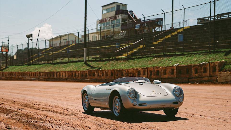 A silver classic Porsche 550 J convertible sports car is parked on a dirt racetrack with empty stadium seating and a building in the background