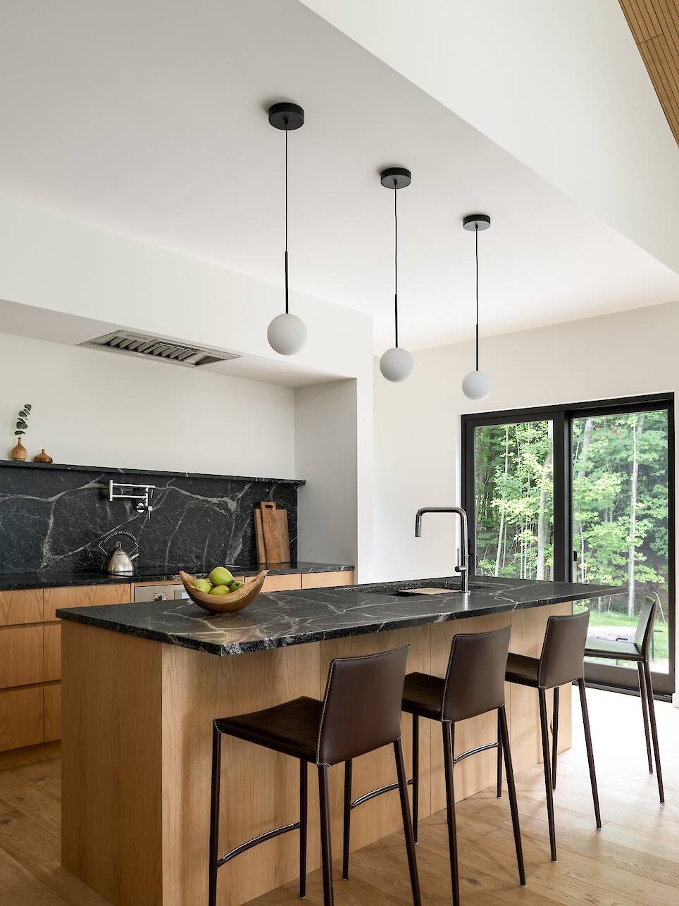 Modern kitchen with wood cabinets, black marble countertops, three pendant lights, a fruit bowl, and three brown bar stools near large glass doors overlooking greenery