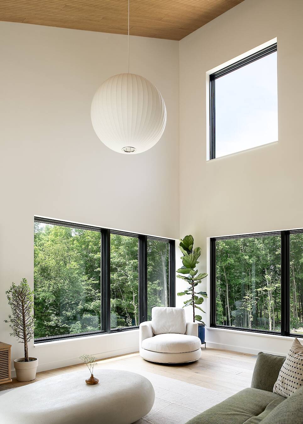 Modern living room with large windows, white walls, a round white chair, green plant, and spherical ceiling light, overlooking a green forest