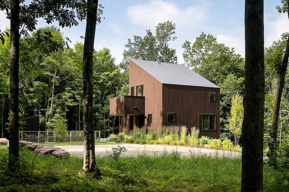 Modern two-story house with dark wood siding and metal roof, surrounded by trees and greenery, with a gravel driveway in front