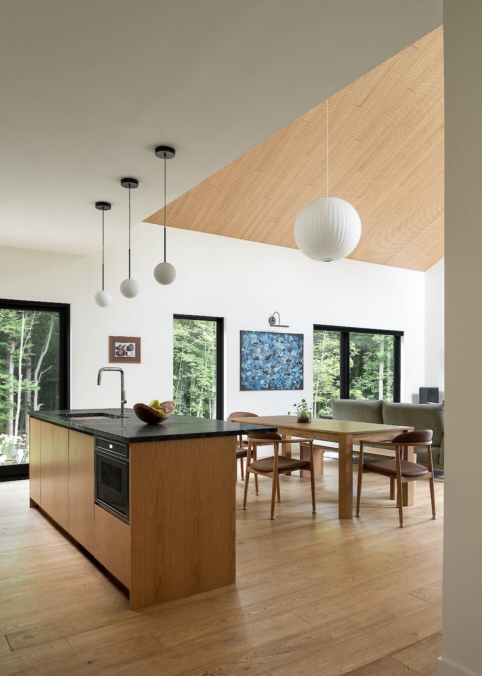 Modern kitchen and dining area with wood floors, large windows, black countertops, pendant lighting, and forest view outside