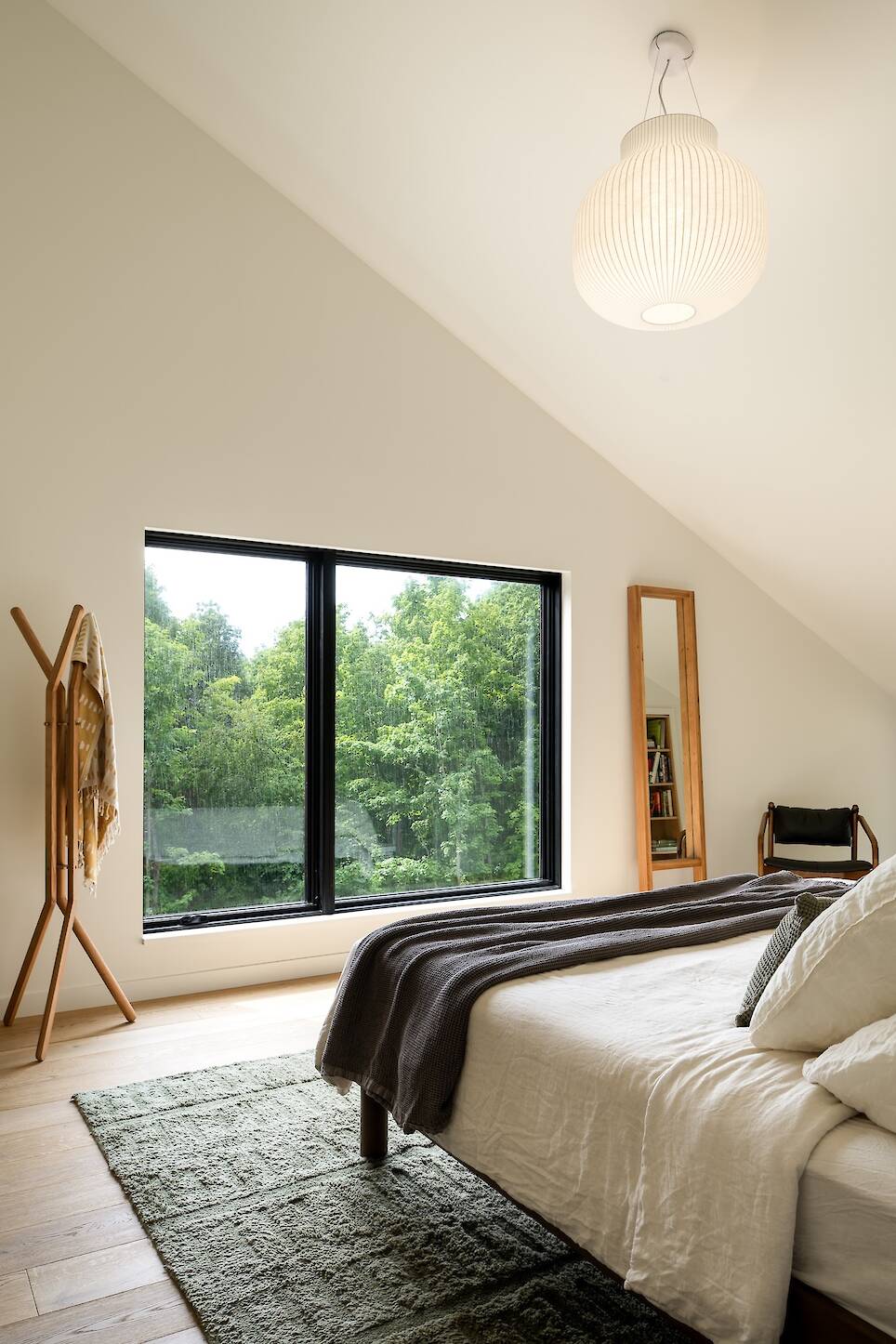 Minimalist bedroom with large window overlooking trees, sloped ceiling, bed with neutral bedding, green rug, wooden coat rack, tall mirror, and chair
