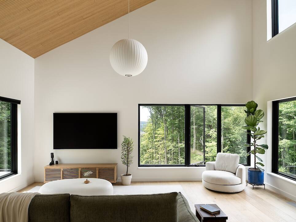 Modern living room with large windows, wall-mounted TV, green sofa, round white chair, potted plants, and light wood ceiling. Natural light fills the space