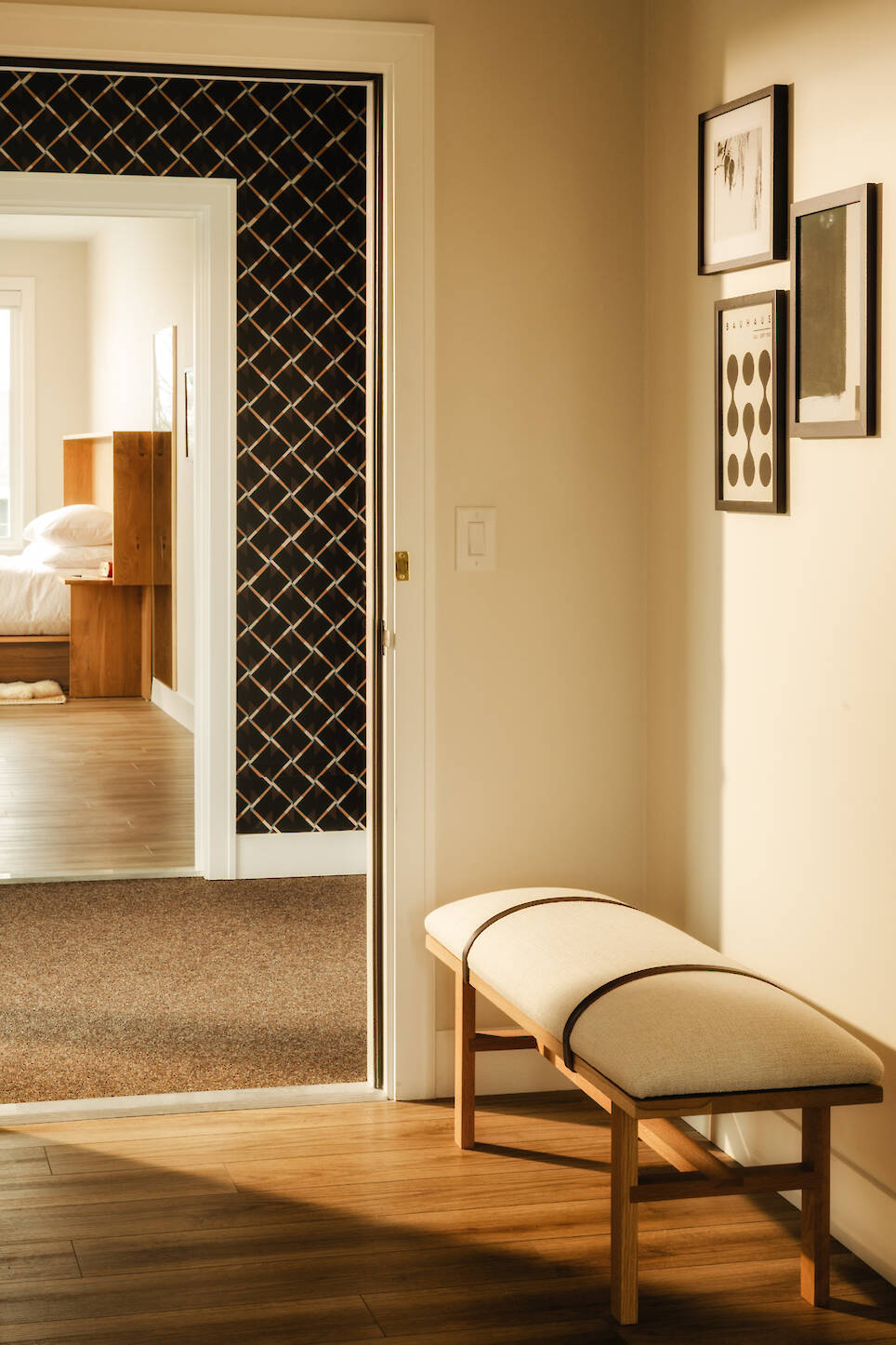 A hallway in the style of the Dexter Hotel Michigan features a cushioned wooden bench, framed art, and an open doorway to a bedroom with wood furniture and a patterned black acc