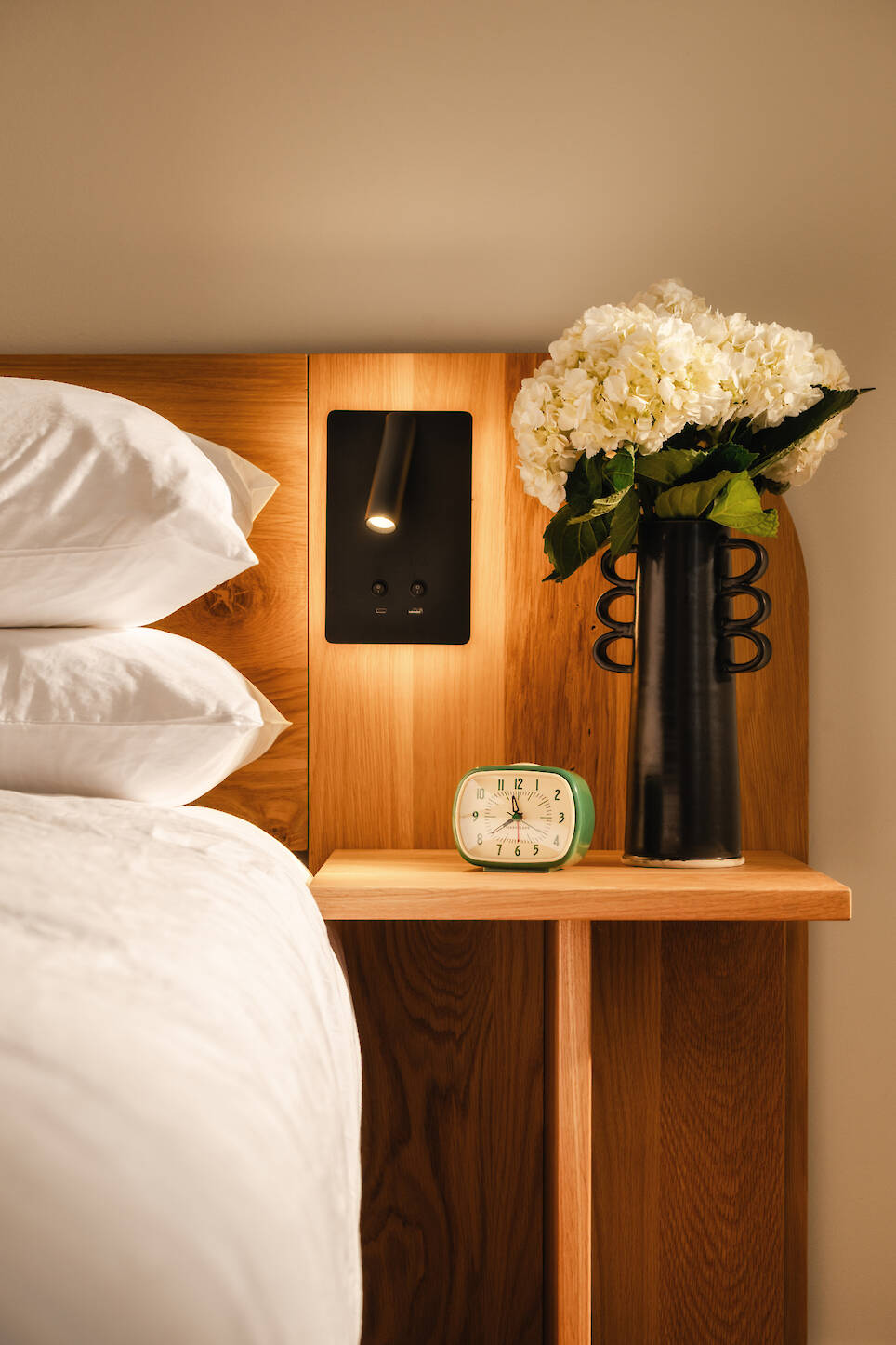 A wooden bedside table at Dexter Hotel Michigan with a black vase of white flowers, a small green alarm clock, wall reading light, and bed with white linens