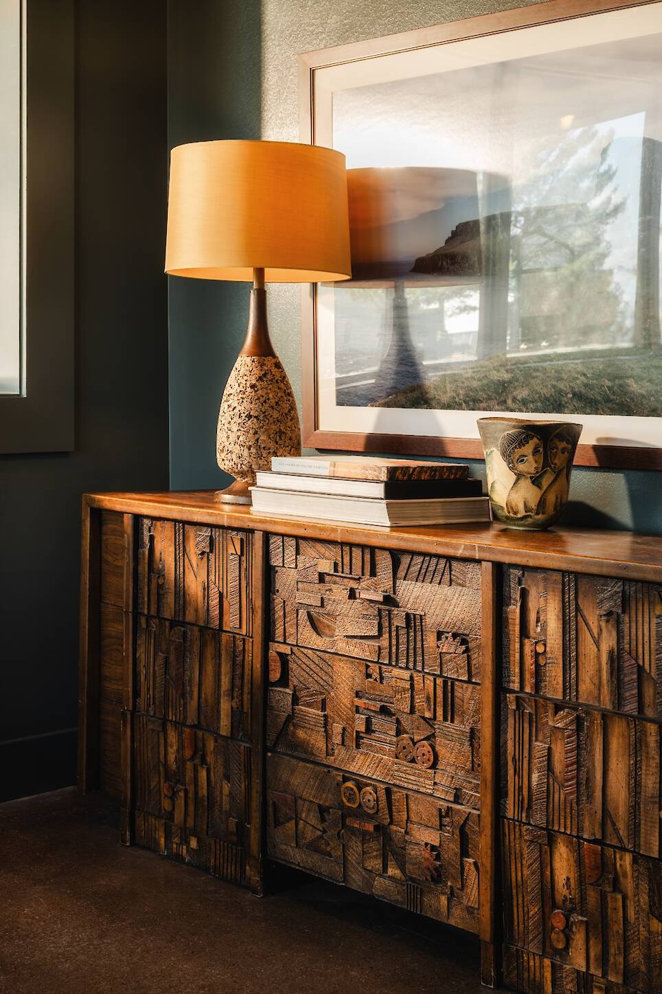 A wooden sideboard with carved geometric patterns, books, a lamp with a beige shade, a decorative bowl, and framed landscape art on the wall behind