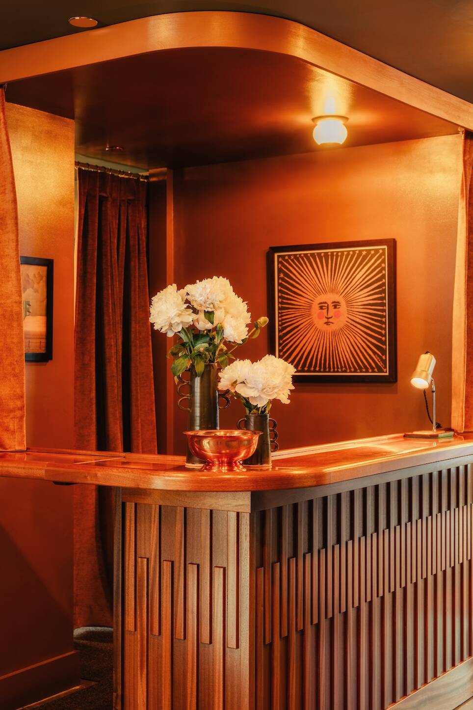 Wooden reception desk with vases of white flowers, copper-colored walls, framed sun artwork, and soft warm lighting