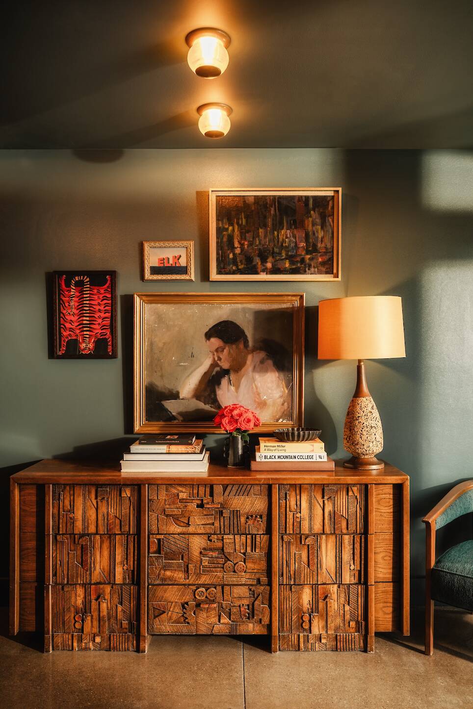 Wooden sideboard with geometric patterns, art and books on top, a table lamp, red flowers, and three framed artworks on a green wall, warmly lit by ceiling lights