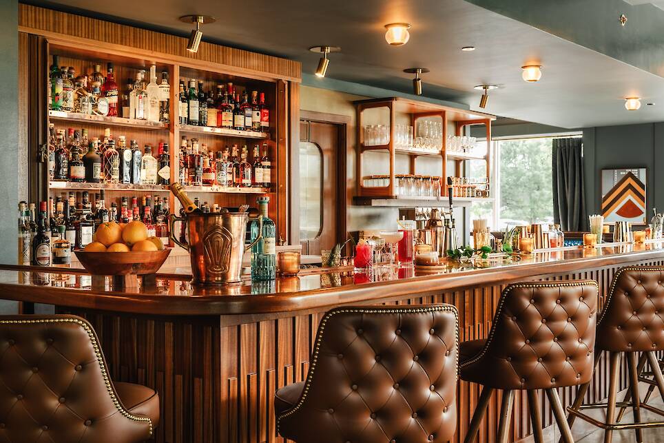A stylish bar with a wooden counter, leather bar stools, and shelves stocked with bottles, glasses, and fruit. Warm lighting creates a cozy atmosphere