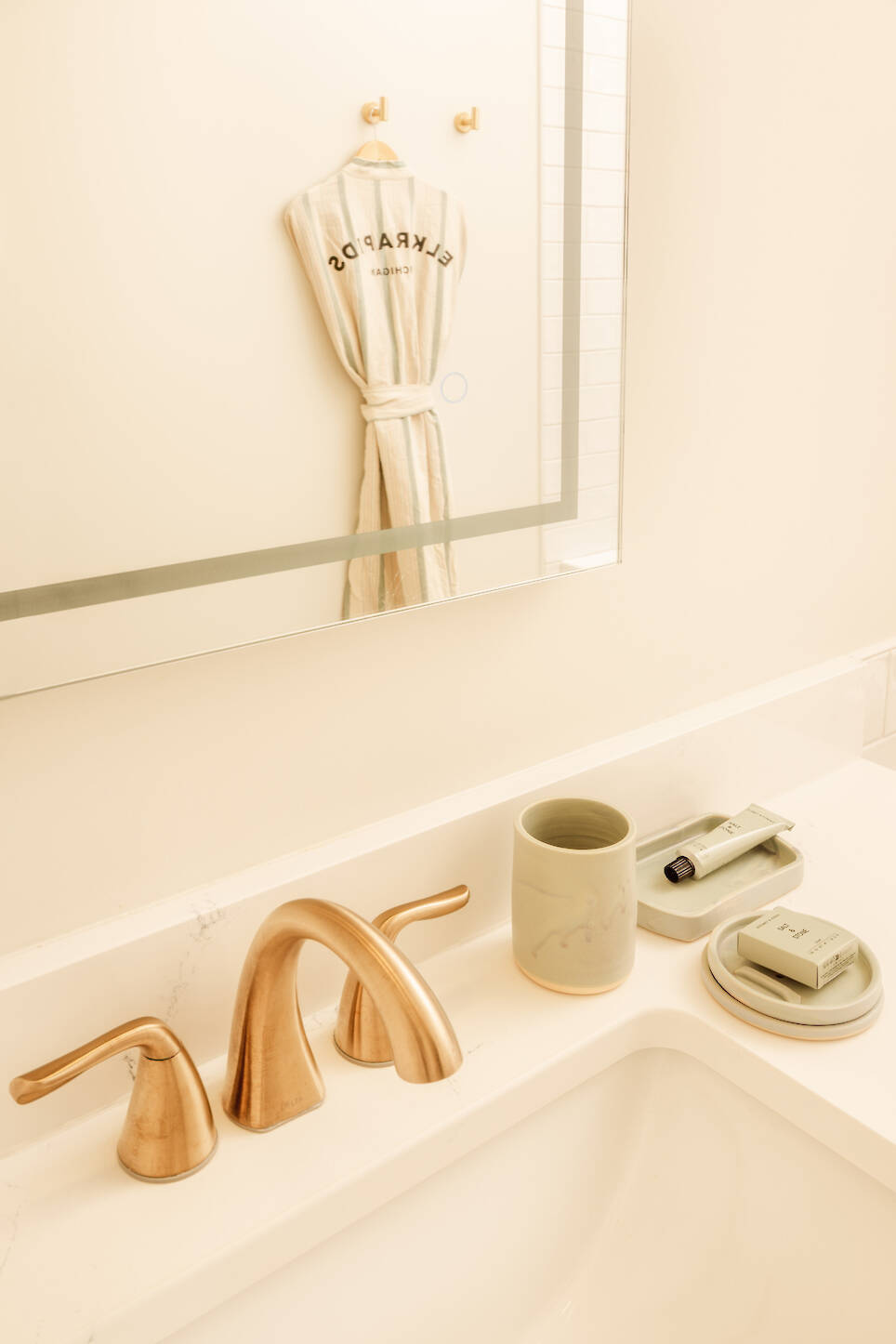 A bathroom sink with gold faucet, soap dish, toothbrush holder, and a robe hanging on the wall reflected in the mirror at Dexter Hotel Michigan