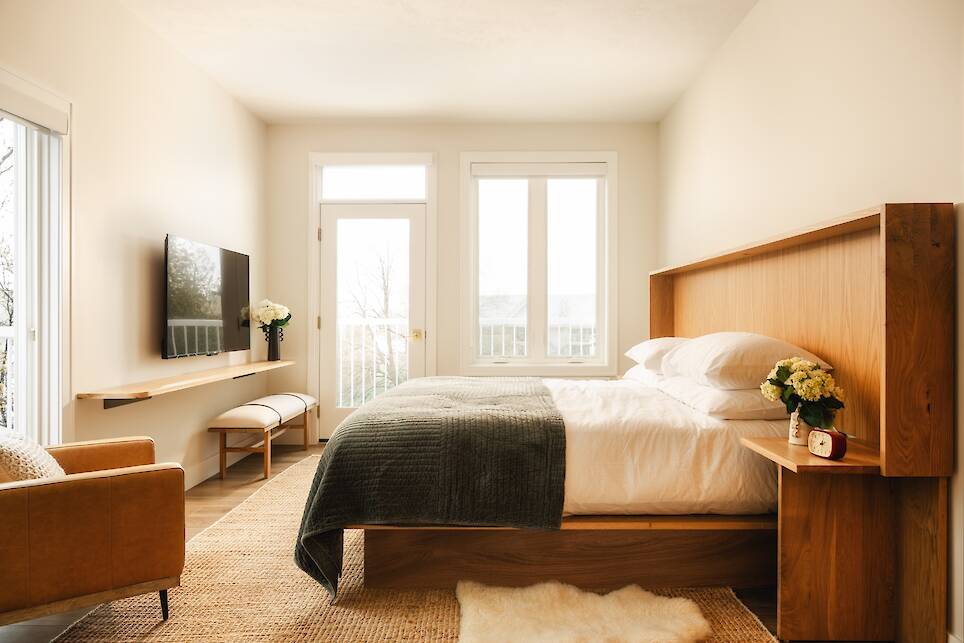 Modern bedroom with a wooden bed, white bedding, green blanket, TV on the wall, tan armchair, flowers, and large windows letting in natural light