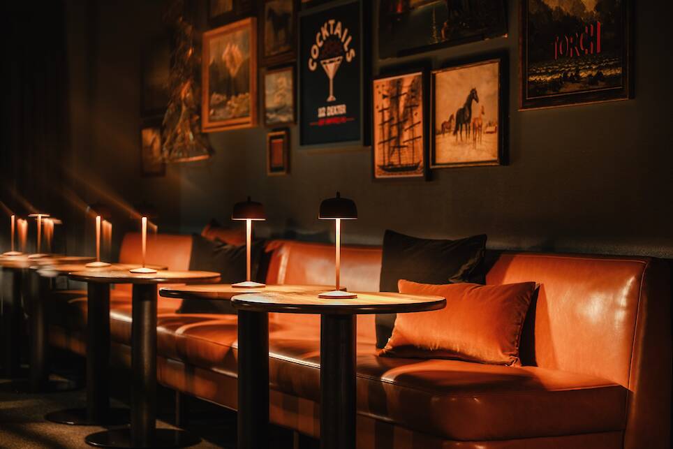 A dimly lit lounge with brown leather seating, small round tables, and wall art, illuminated by modern table lamps