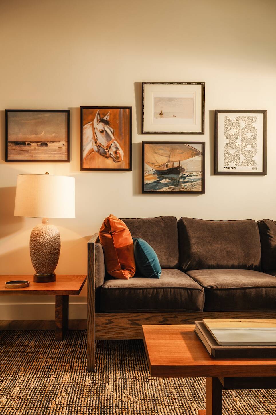 A modern living room with a dark sofa, orange and blue cushions, a lamp on a wooden side table, and framed artwork on the wall above