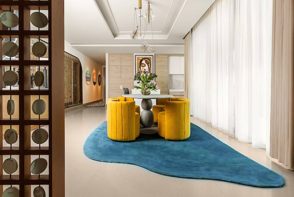 Modern dining area with yellow chairs around a white table on a blue rug, large windows with sheer curtains, and wall art in the background
