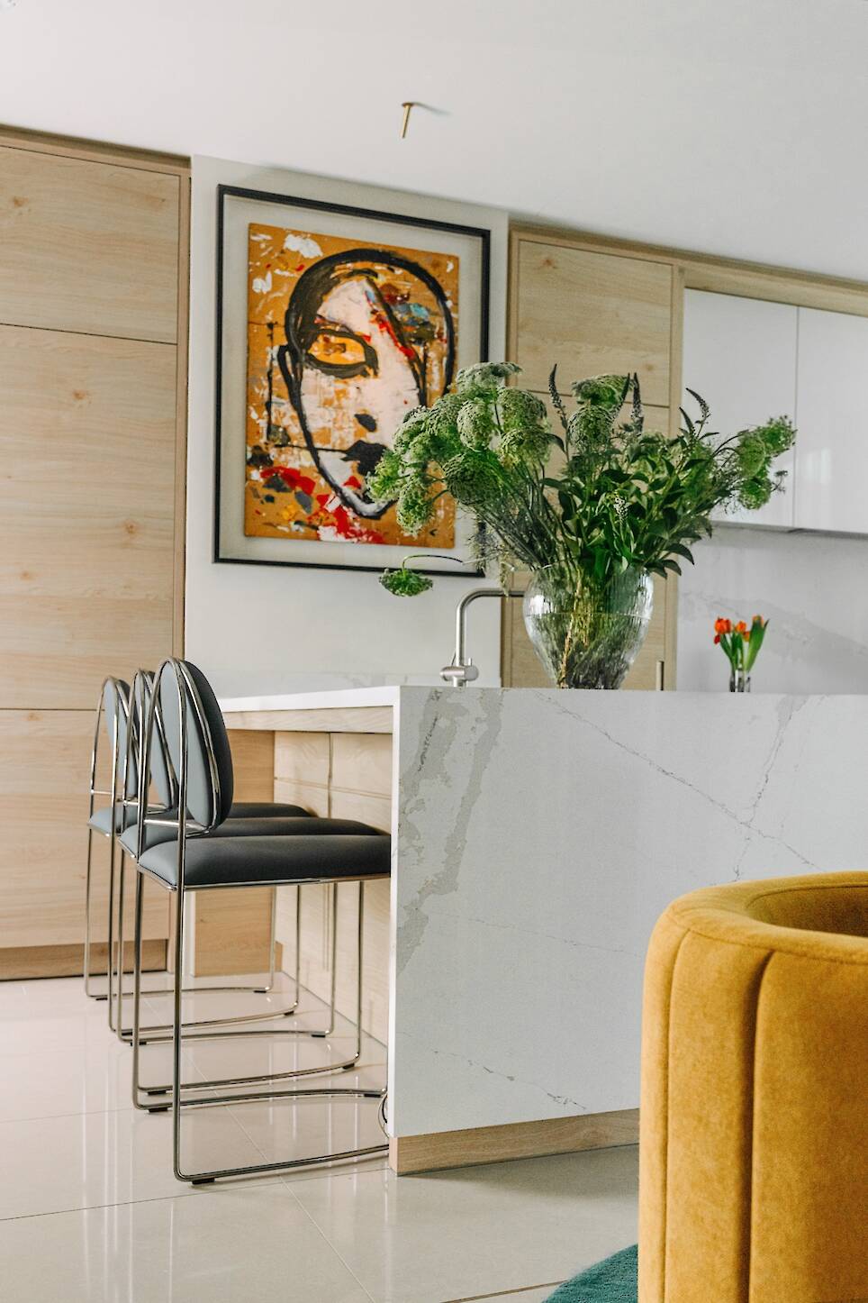 Modern kitchen with a marble island, black chairs, a large vase of greenery, abstract face artwork on the wall, and light wood cabinetry