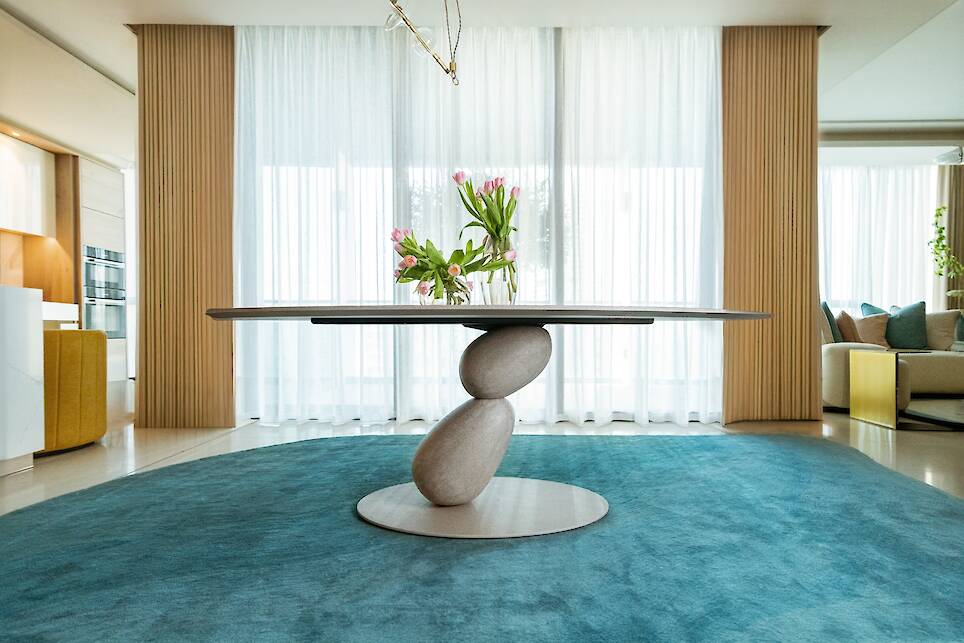 Modern dining room with a sculptural stone-based table, a vase of pink tulips on top, blue circular rug, and sheer curtains allowing natural light into the space