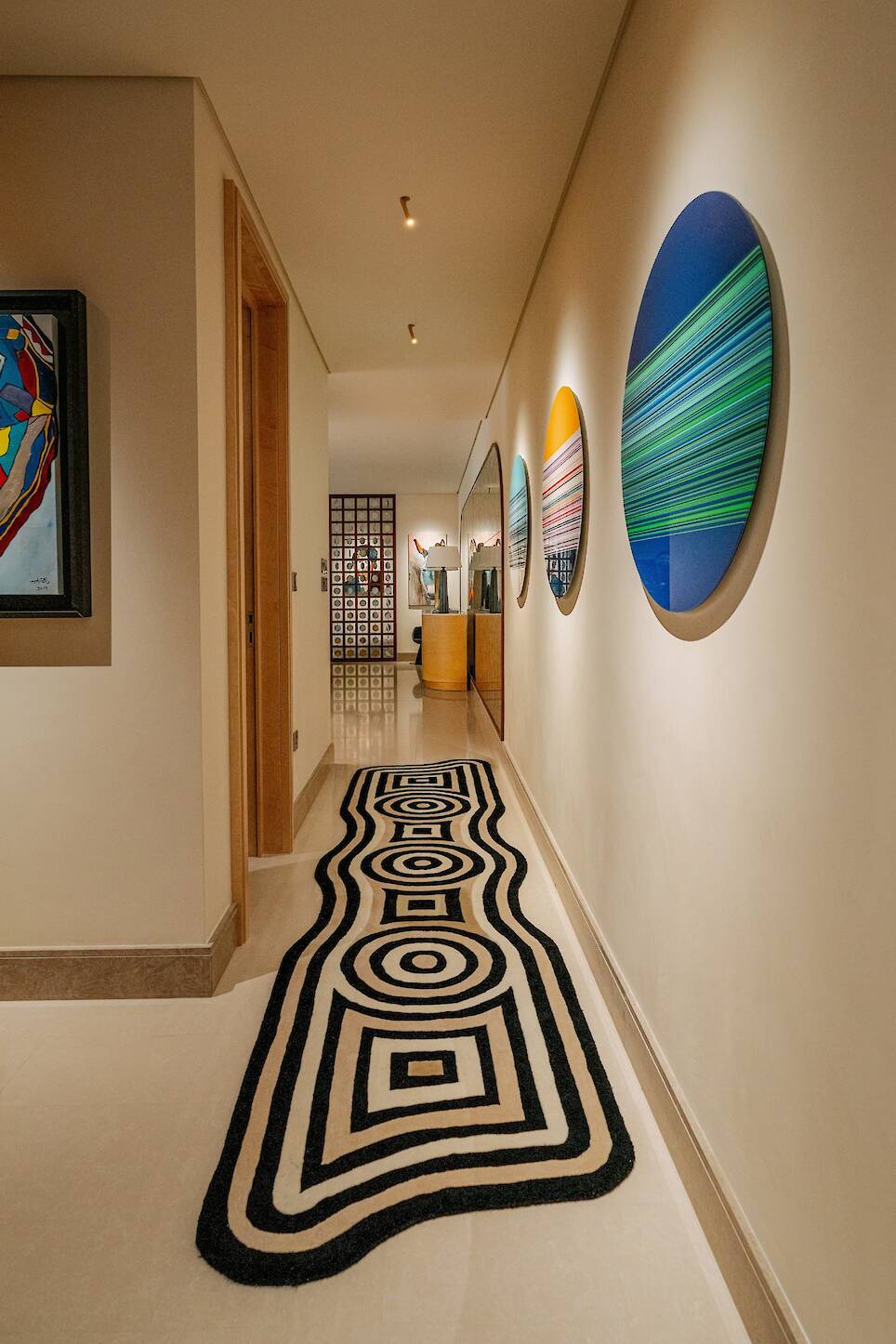 A hallway with a patterned black and white rug, abstract circular art on the walls, and neutral-toned decor leading to a distant room