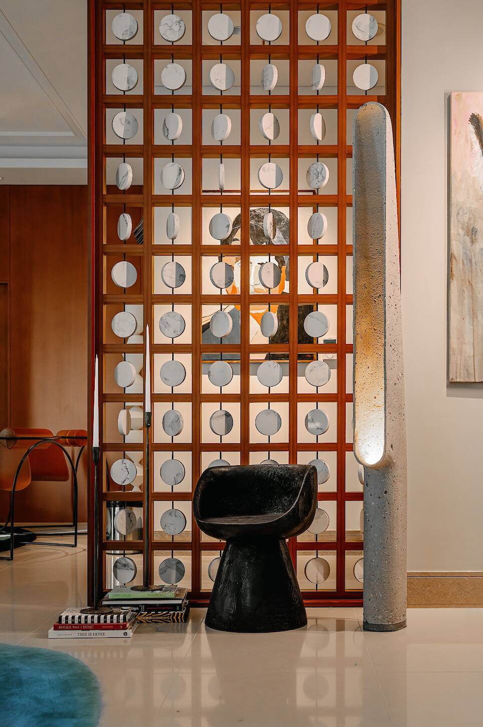 Modern interior with a black sculptural chair, tall concrete floor lamp, books on the floor, and a decorative wooden partition with circular accents. Abstract artwork is visible