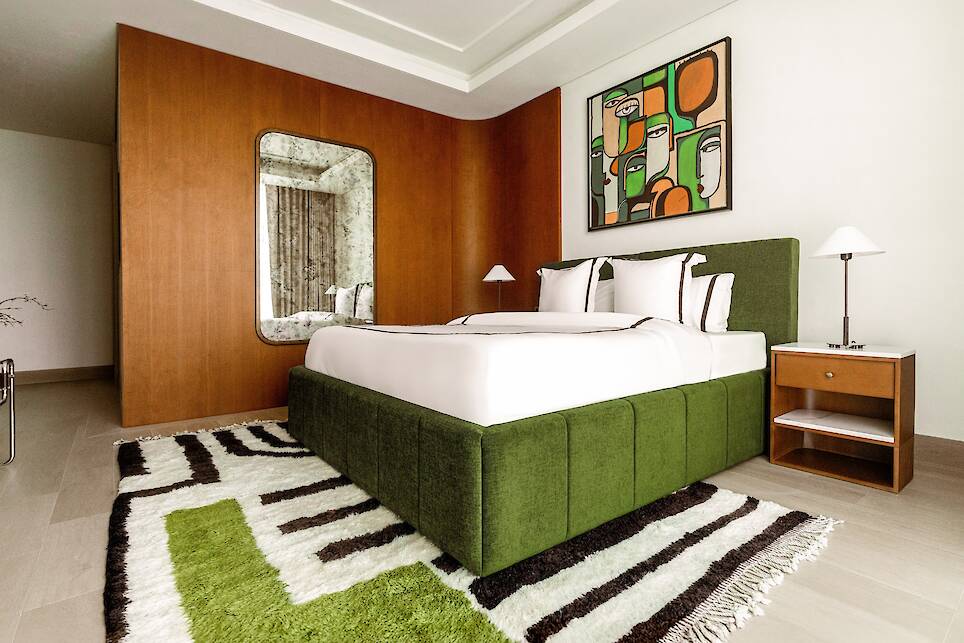 Modern bedroom with a green upholstered bed, geometric rug, wooden wall with a mirror, abstract art above bed, and two nightstands with lamps
