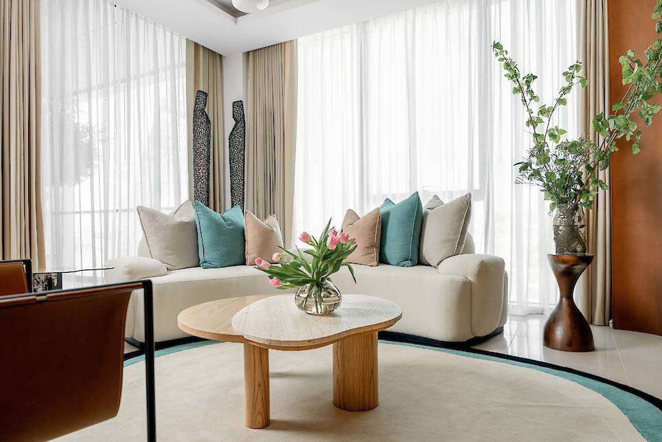 Modern living room with a curved white sofa, assorted pillows, wooden coffee table with pink tulips, large windows with sheer curtains, and plants on the side