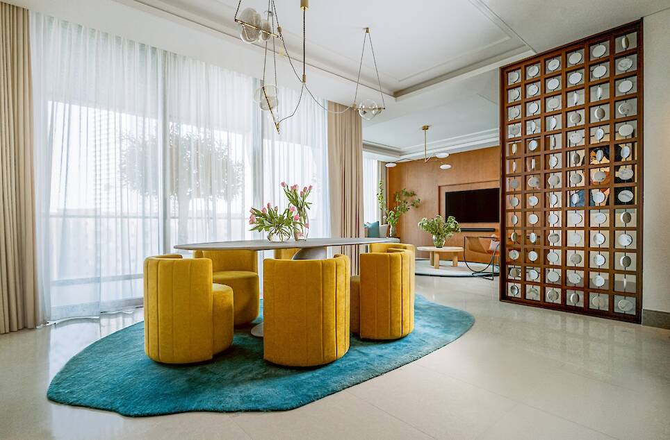 Modern dining area with a round table, six yellow chairs on a blue rug, large windows with sheer curtains, and a partition separating the living room with a TV and plants