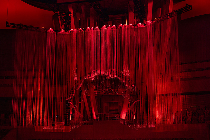 a frontal view of the hanging structure lit deep red, with the concert hall’s organ visible through the strands