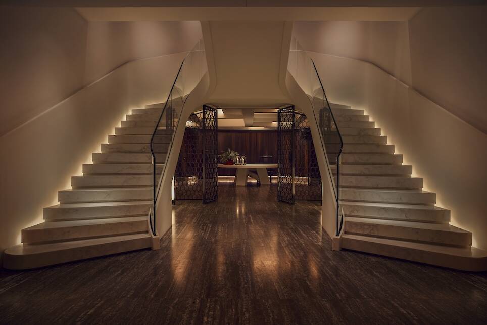 A grand staircase with illuminated steps curves upward on both sides of a polished hallway, leading to an upper level