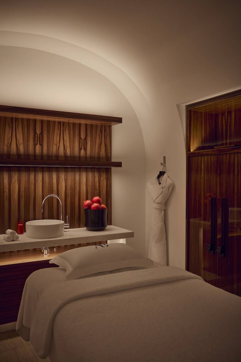 A spa room with a massage table, sink, robe on a hook, bowl of red apples, wooden cabinets, and warm ambient lighting