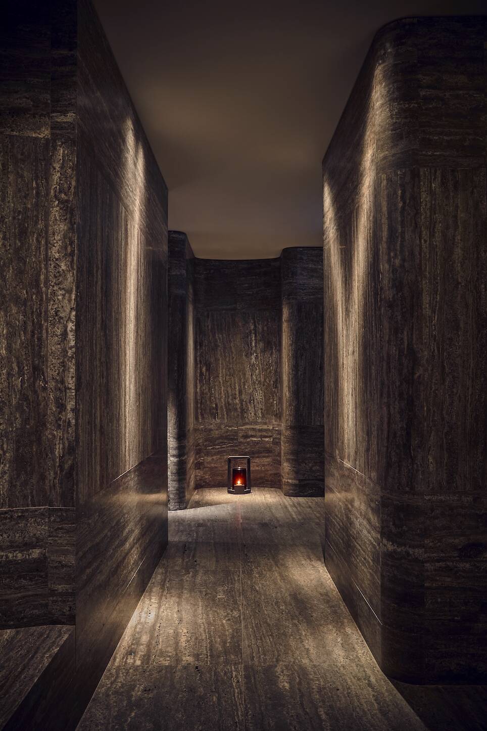 Dimly lit corridor with smooth, dark stone walls and floor, featuring a small candle lantern at the end of the hallway
