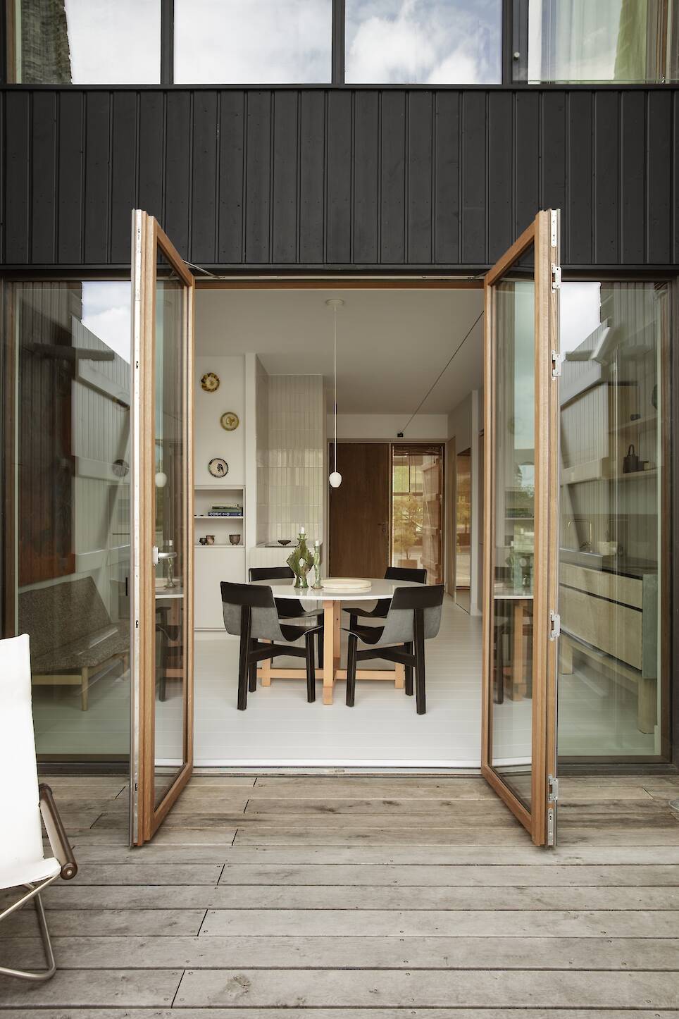 Open glass doors lead from a wooden patio into a modern dining area with a table, four chairs, and minimal decor on the walls