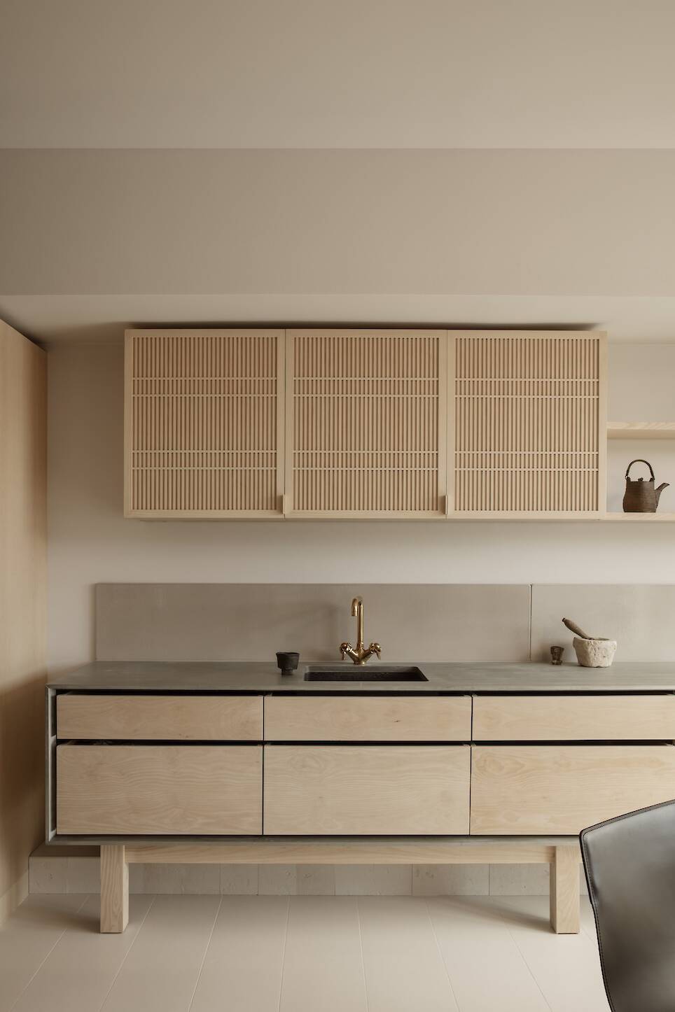 Minimalist kitchen with light wood cabinets, a black countertop, gold faucet, and neutral walls. Upper cabinets have slatted doors; a small shelf holds decorative items