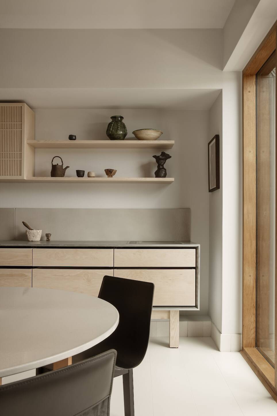Minimalist kitchen with light wood cabinets, open shelves displaying ceramics, a round table, black chairs, and large window letting in natural light