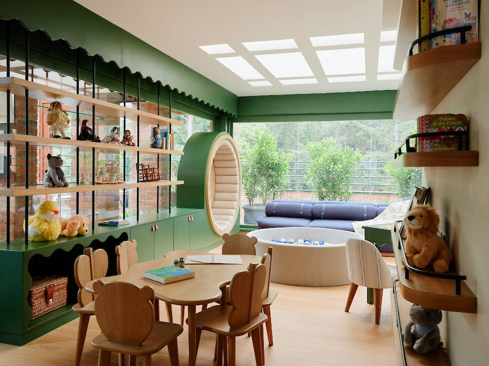 Children’s playroom with wooden tables and bear-shaped chairs, a circular reading nook, a ball pit, shelves filled with toys and books, and a view of trees through large windows
